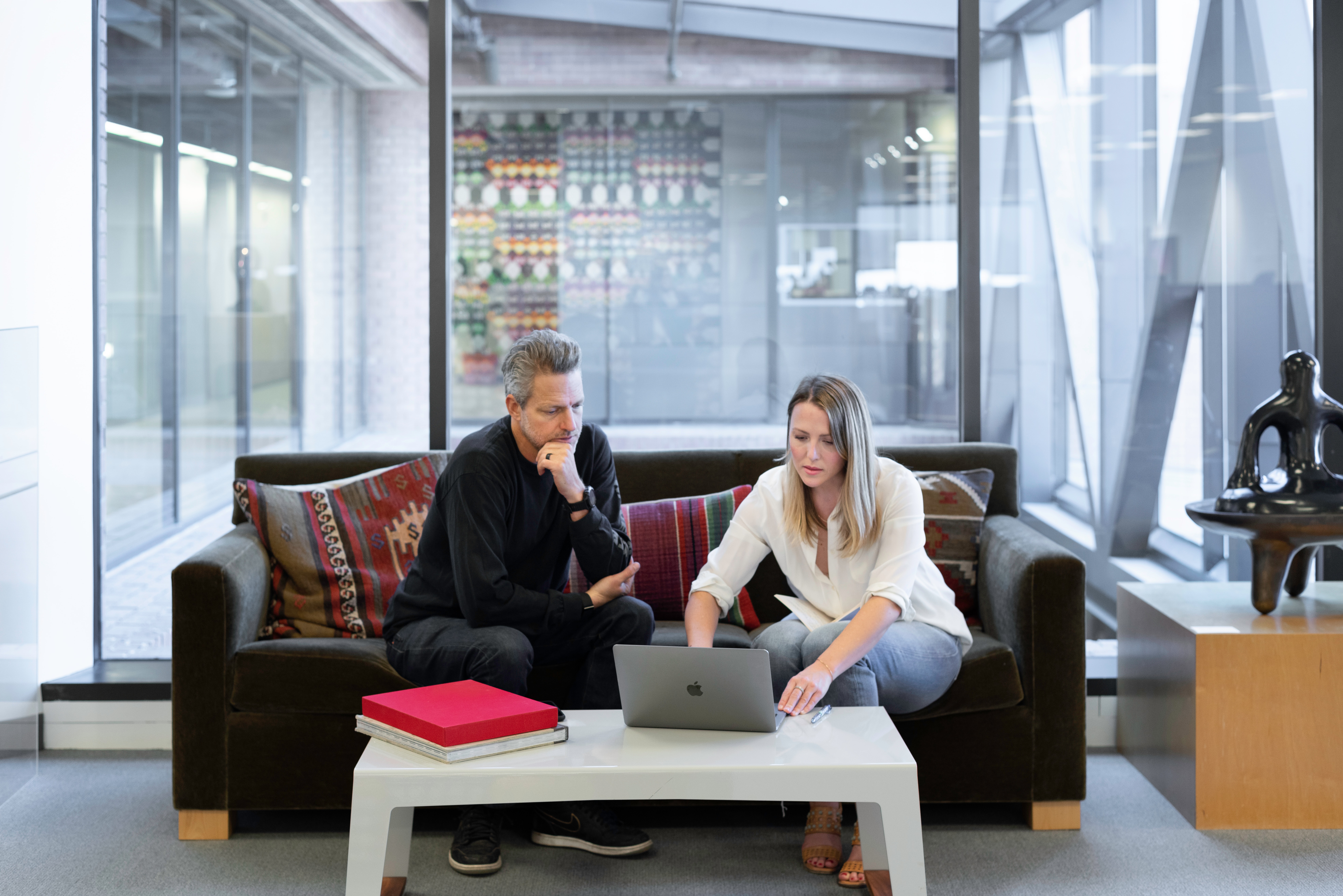 Man and women looking at laptop on sofa with glass office behind